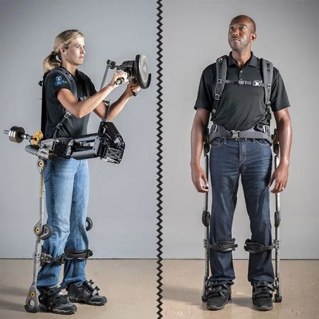 Two people wearing heavy industrial exoskeletons stand in a grey studio.