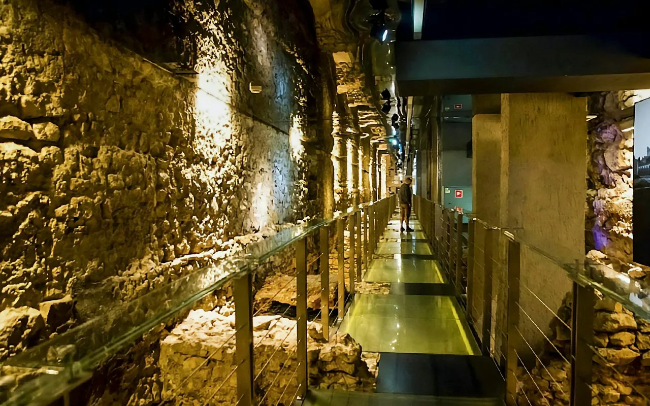 A lone figure walks a modern walkway through ancient, illuminated underground ruins.