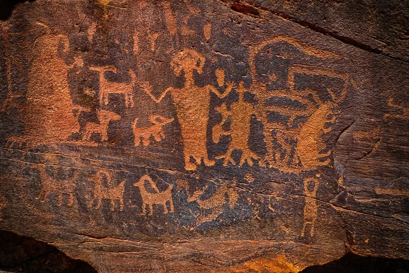 Ancient petroglyphs carved into reddish-brown rock, depicting stylized figures and animals.