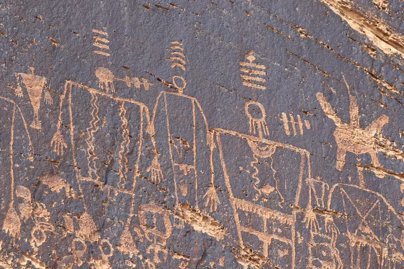 Ancient petroglyphs carved into dark rock showing humanoid figures and symbols.